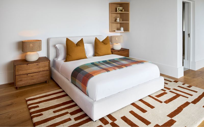 A geometric hand-tufted bedroom floor rug in rich rust and burnt orange, placed in a partial layout beneath a white bed on wooden flooring.